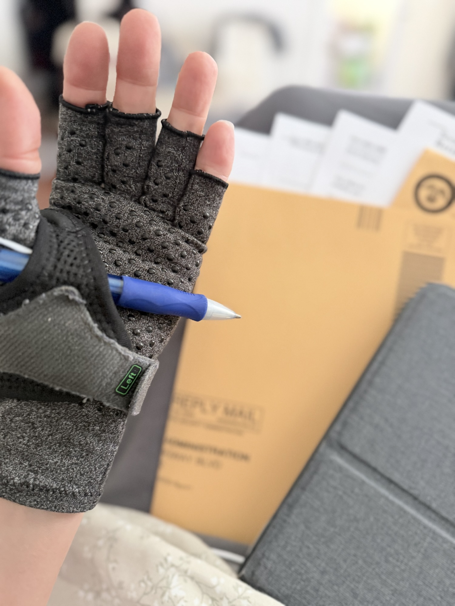 Michelle’s left hand in a gray brace, with a blue pen tucked across her palm, in front of disability application paperwork and a mailing envelope.